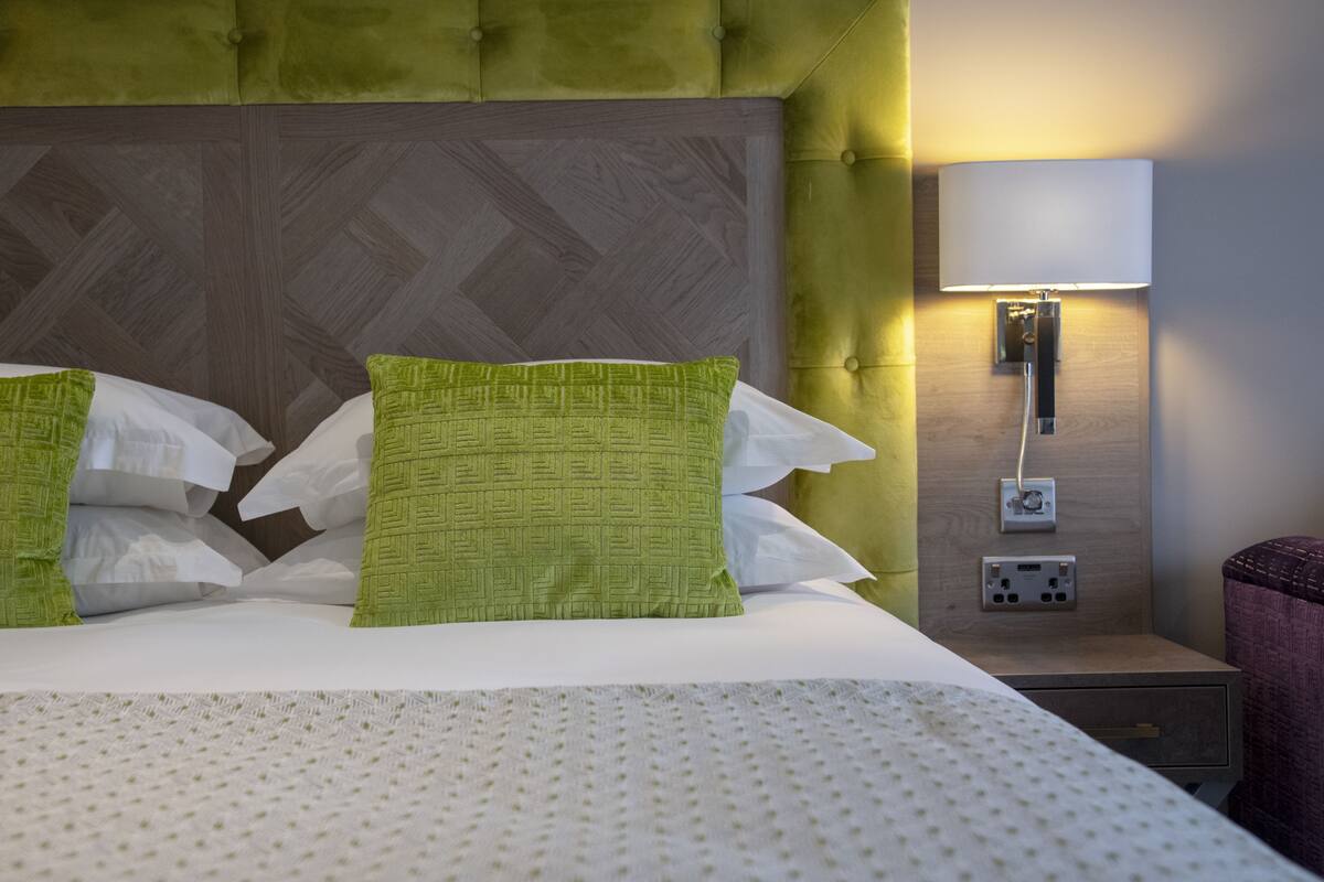 A neatly made bed with a patterned bedspread, two green pillows, a wooden headboard, and a bedside lamp.