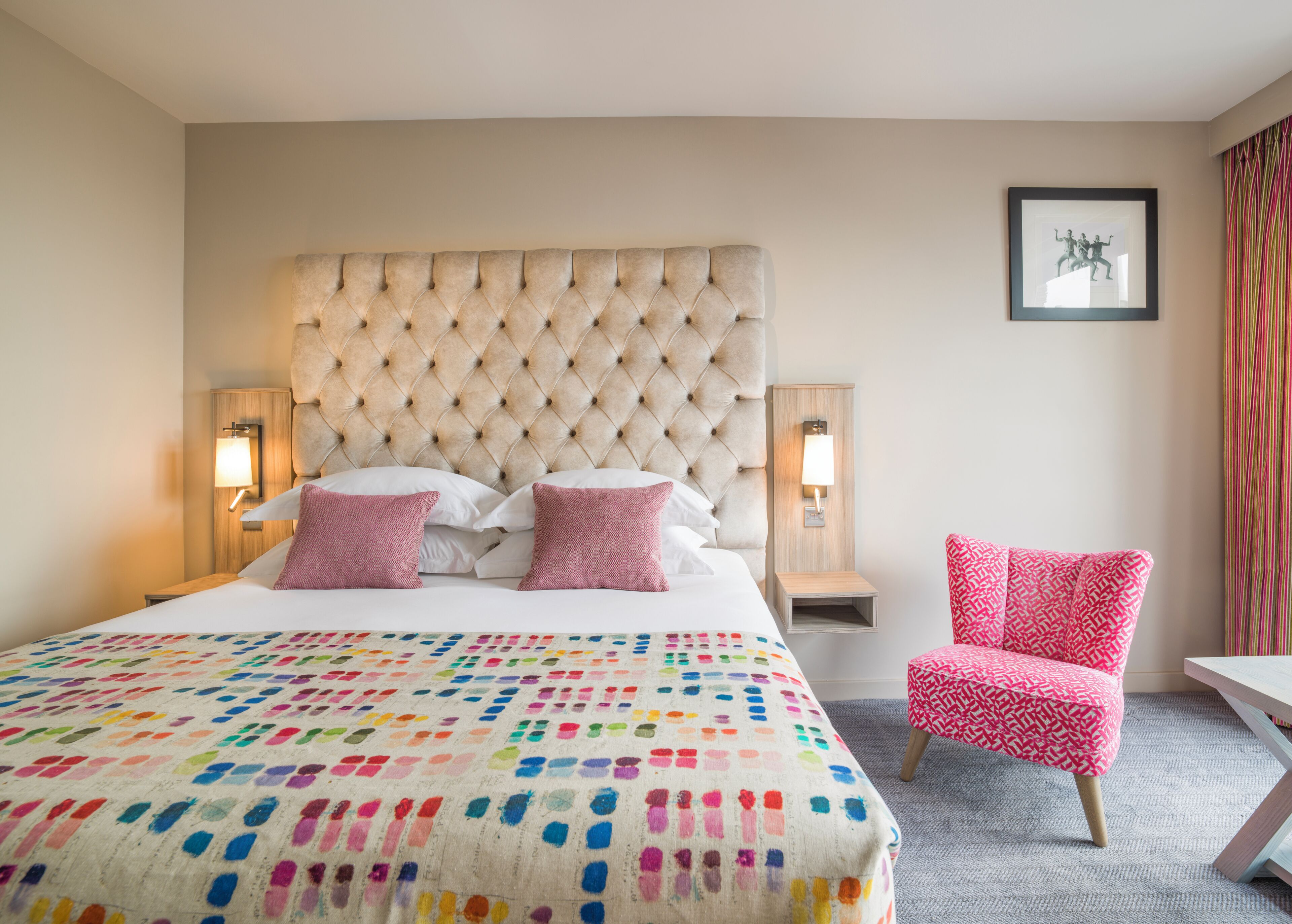 Une chambre d’hôtel avec une tête de lit capitonnée, une couvre-lit aux couleurs vives, un fauteuil à motifs roses et un tableau encadré au mur.