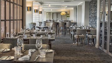 A spacious hotel dining area with multiple tables set for guests, featuring a modern design with neutral tones and a patterned carpet.