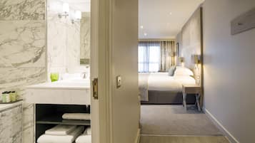 A modern hotel room with a marble bathroom, a neatly made bed, and a bedside table with a lamp.