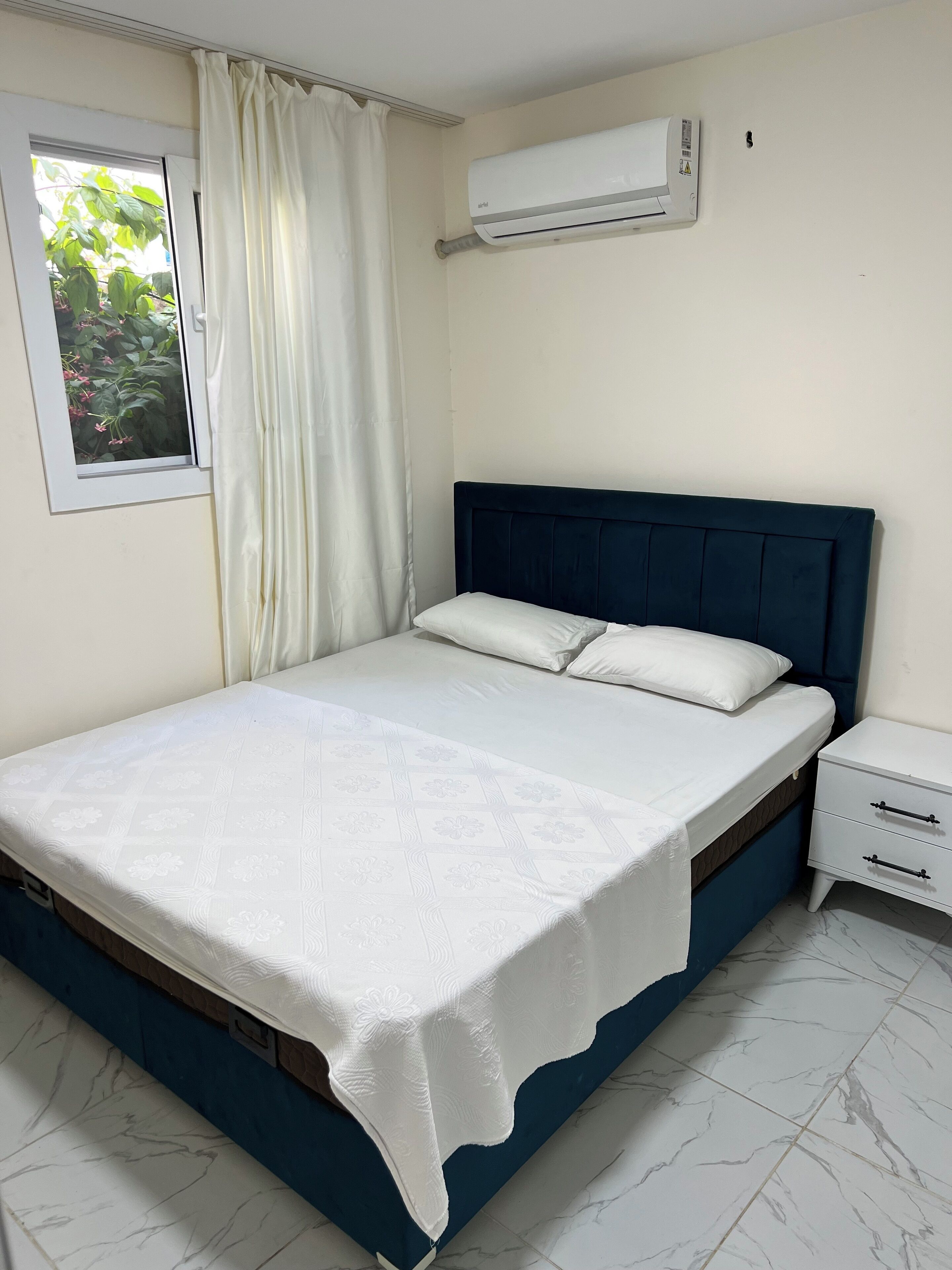 Un dormitorio con cama, mesita de noche, equipo de aire acondicionado, ventana con vista a vegetación y cortina.