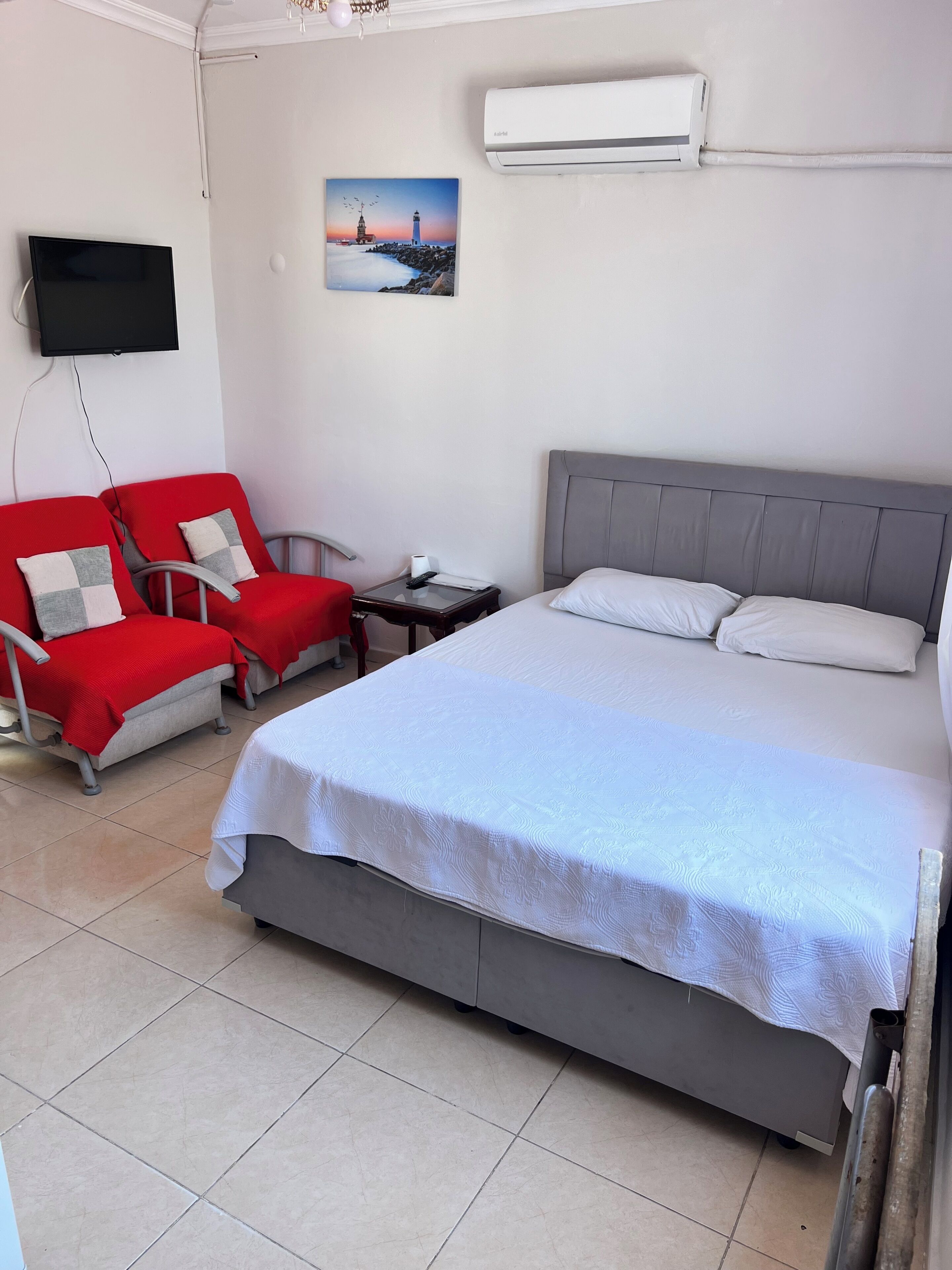 Una habitación de hotel con una cama, televisión, una silla roja, mesita de noche y un cuadro de un faro.