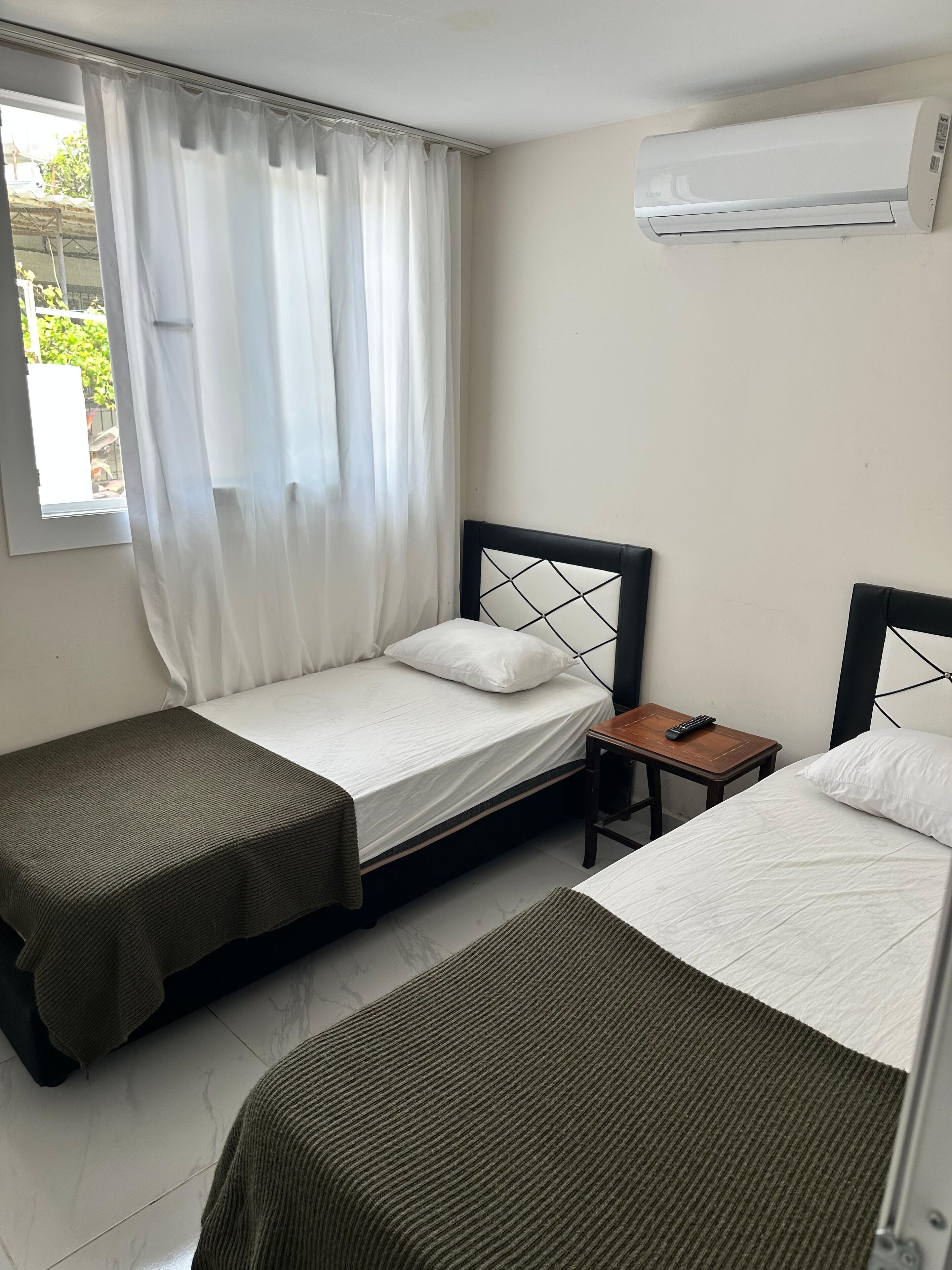Ein Hotelzimmer mit zwei Einzelbetten, einem kleinen Nachttisch aus Holz, einer Klimaanlage und einem Fenster mit weißen Vorhängen.