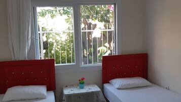 A small, clean room with two single beds, a small table with a vase, and a window with a view of greenery.