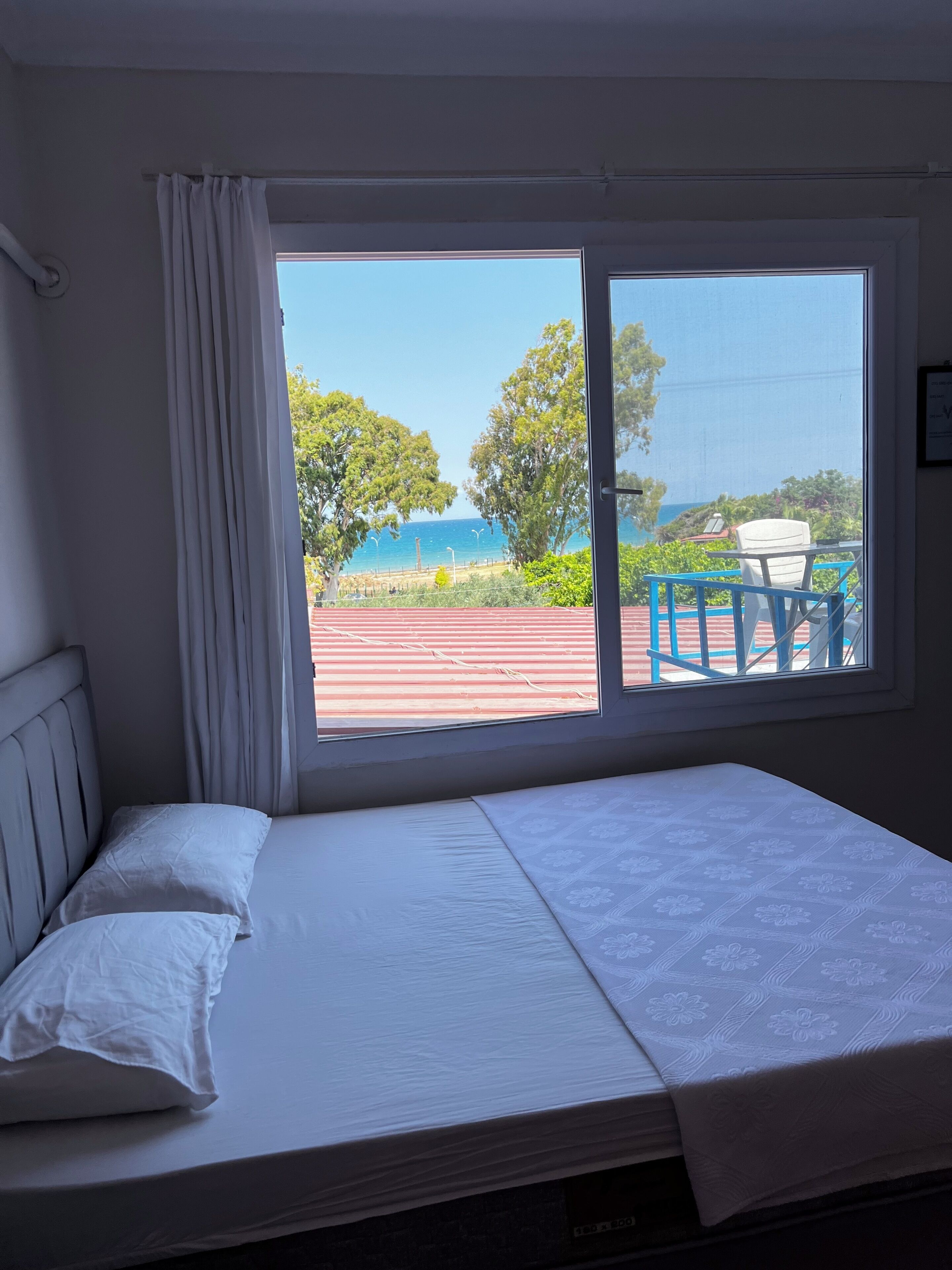 Una habitación con una cama, un ventanal con vista a la playa y árboles, y una silla afuera.