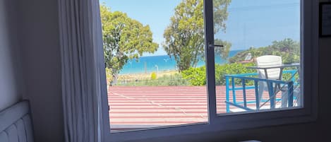 A room with a bed, a window with a view of a beach and trees, and a chair outside.