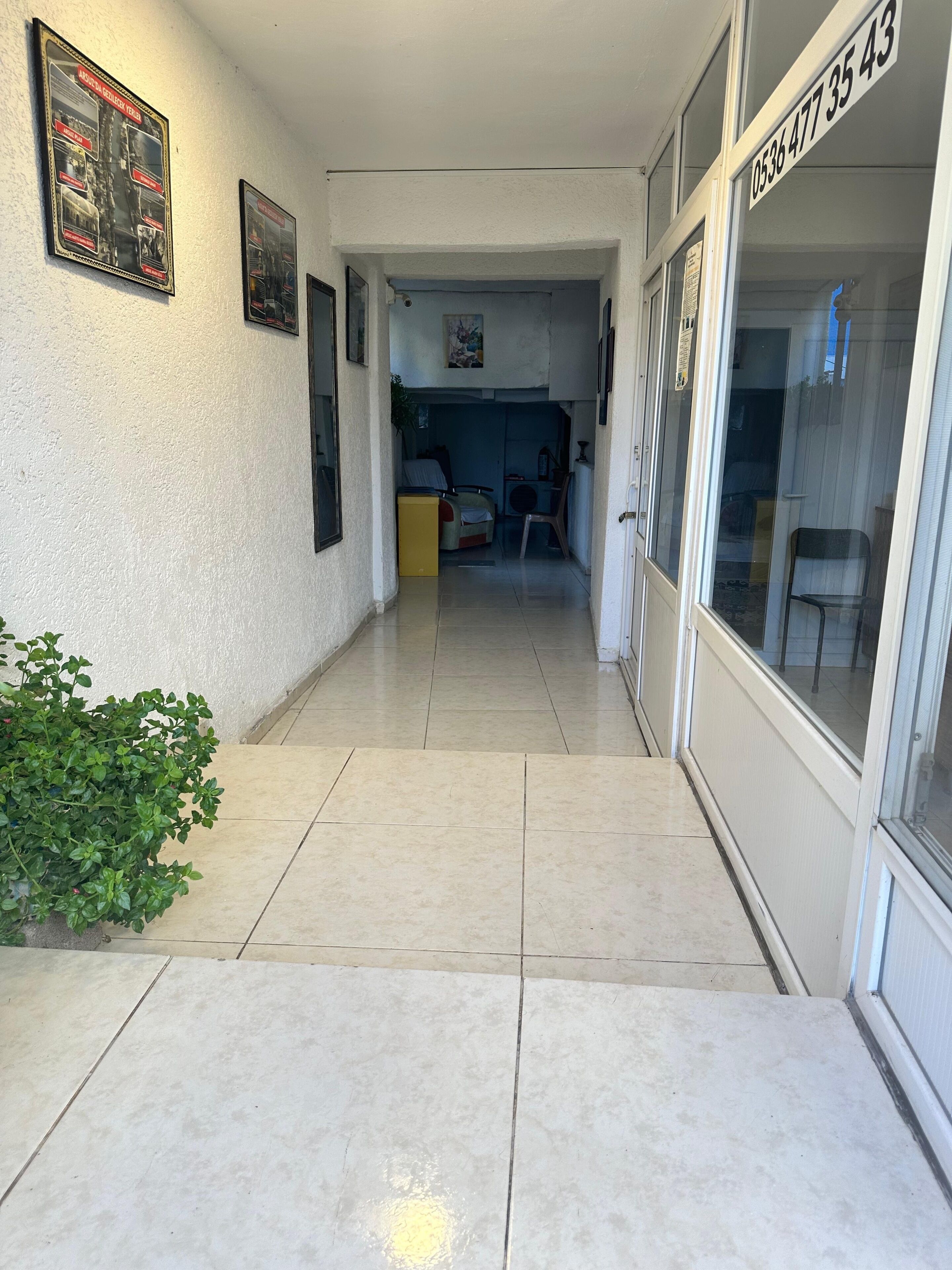 A corridor with tiled flooring, a potted plant, framed pictures on the wall, and a door with a number sign.