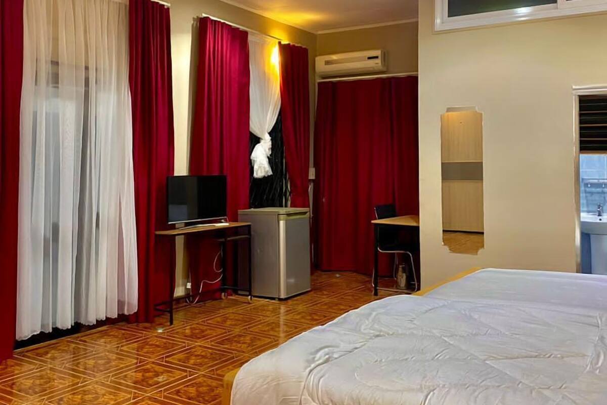 A hotel room with a bed, a desk with a computer, a TV, an air conditioning unit, and a ceiling fan.
