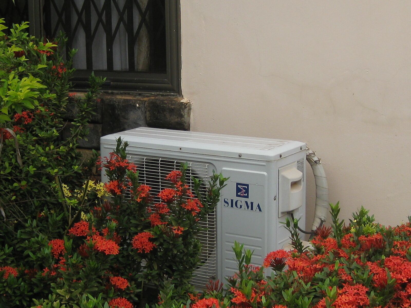 Un appareil de climatisation portant l’inscription « SIGMA » est installé devant un bâtiment doté d’une fenêtre et de plantes en fleurs.