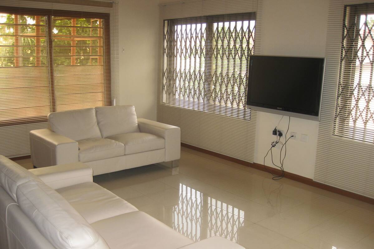 A living room with a white sofa, a flat-screen TV mounted on the wall, and windows with blinds.