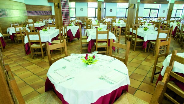 Un salón de banquetes con mesas redondas dispuestas para el servicio de comidas, sillas de madera y suelo de baldosas.