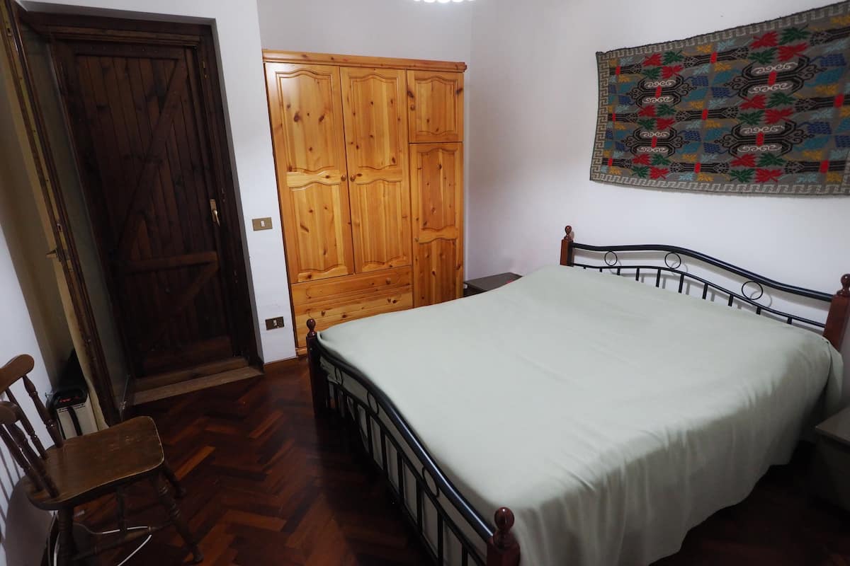 A bedroom with a wooden wardrobe, a bed with a white bedspread, a wooden chair, and a wall tapestry.