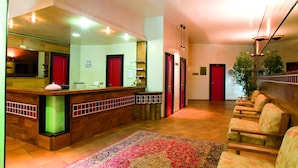 A reception area with a wooden reception desk, red doors, a patterned rug, and a row of upholstered chairs. - Magara Hotel (Spezzano della Sila)