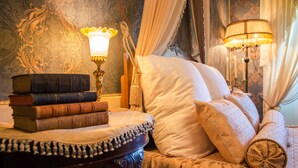 Habitación clásica con papel tapiz decorativo, una cama con ropa de cama blanca y dorada, una mesita de noche con libros y una lámpara.