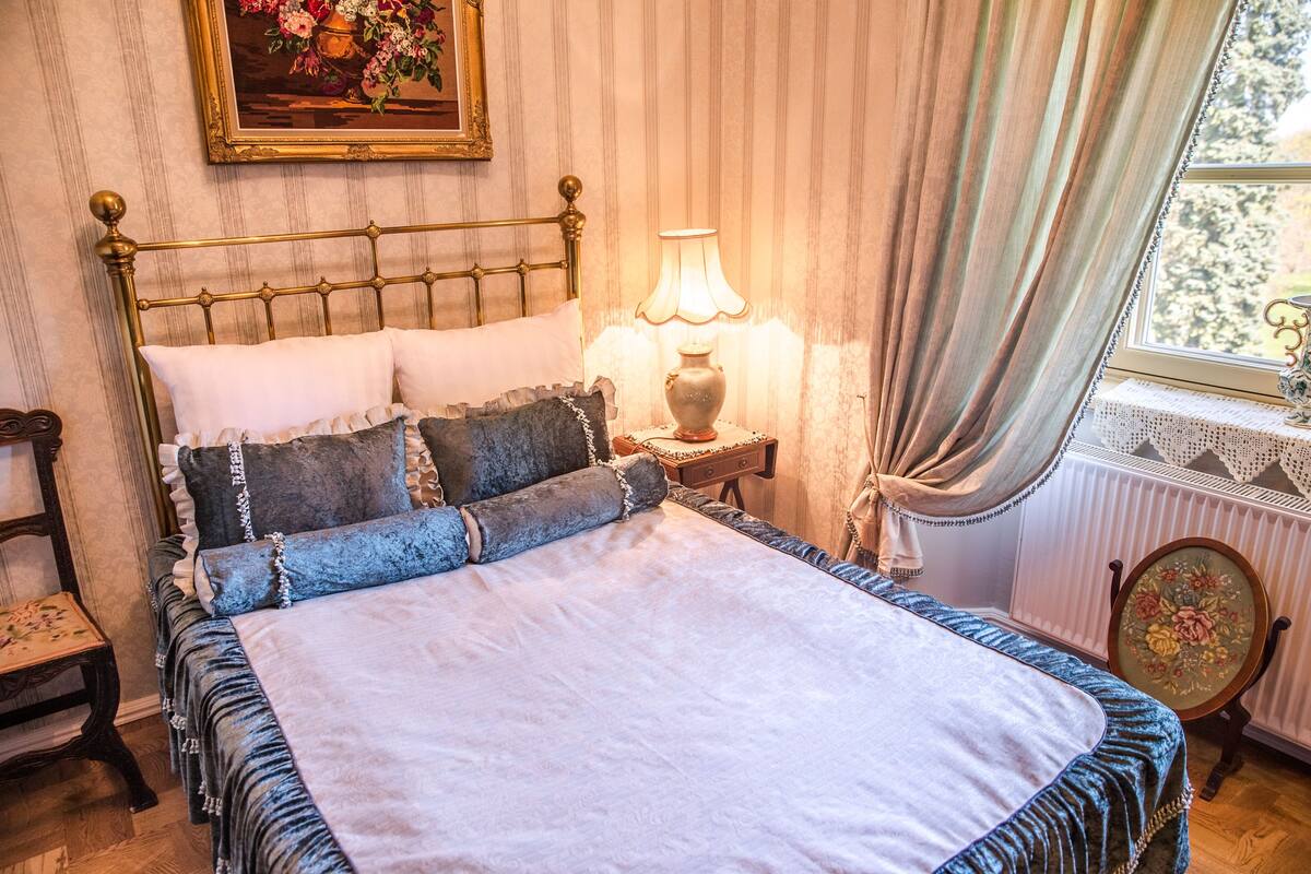 A classic bedroom with a four-poster bed, a bedside table with a lamp, and a window with curtains.