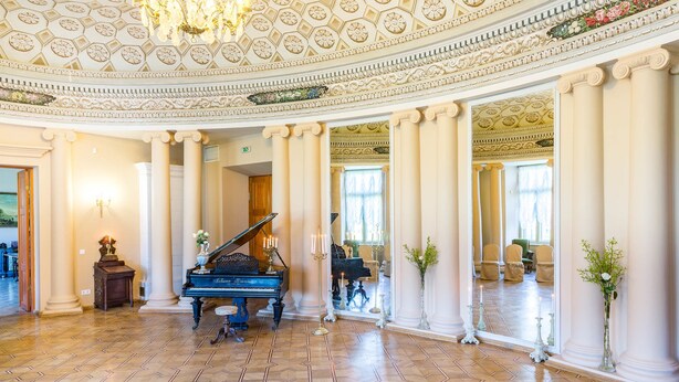 Un salón imponente con un piano de cola, techo ricamente decorado y columnas.