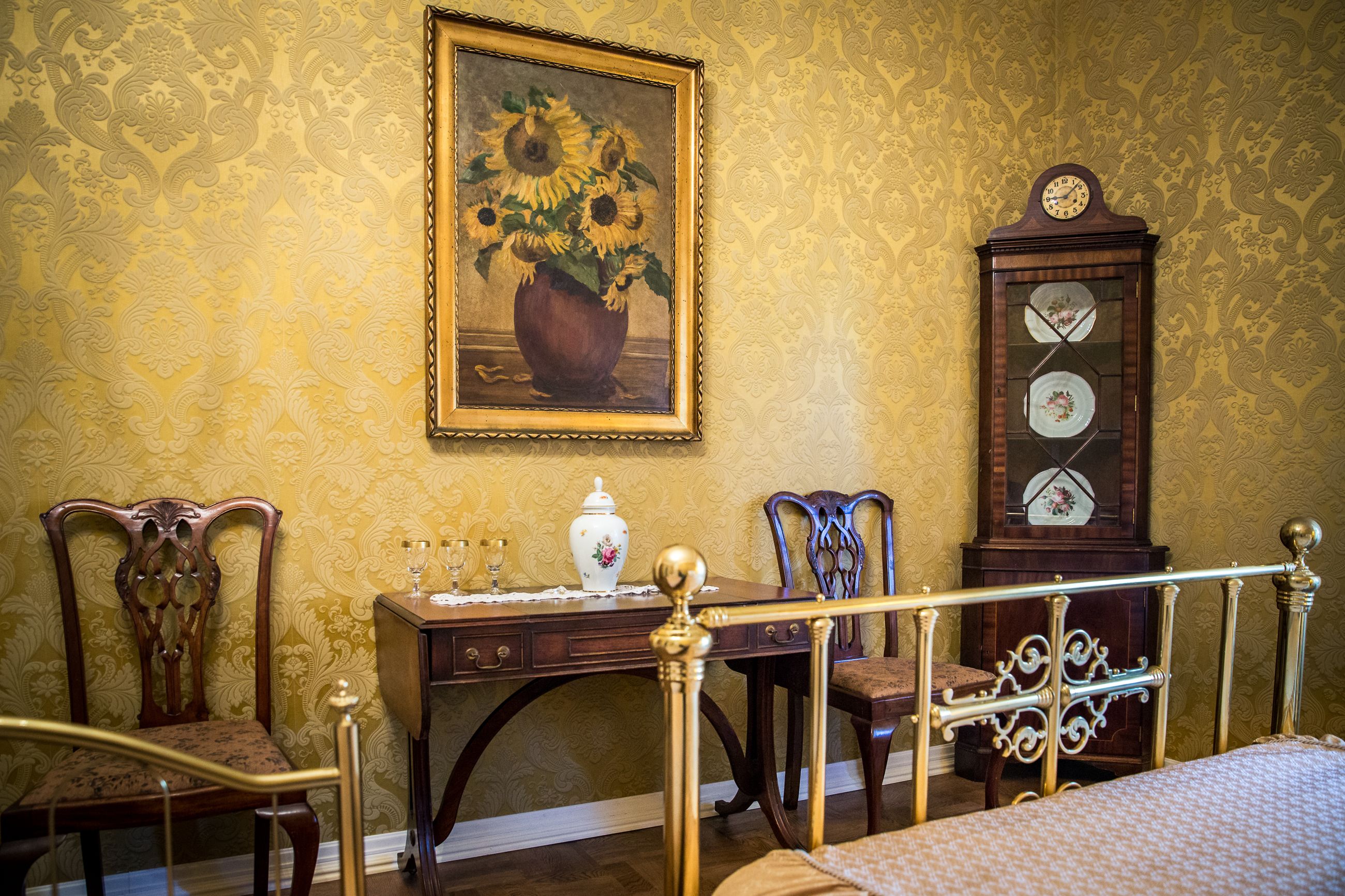 A room with a bed, a wooden desk with a vase and glasses, a chair, and a wall clock.