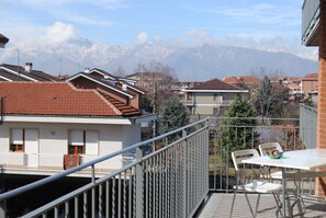 Un balcón con una mesa y sillas, con vista a una zona residencial con casas y una cadena montañosa al fondo.