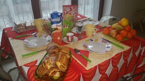 Una mesa de comedor dispuesta con platos, cubiertos, un recipiente de vidrio con jugo de naranja, una botella de agua, una cesta de naranjas, un vaso de agua, un vaso de jugo y un plato con pasteles, junto con una tarjeta decorativa.