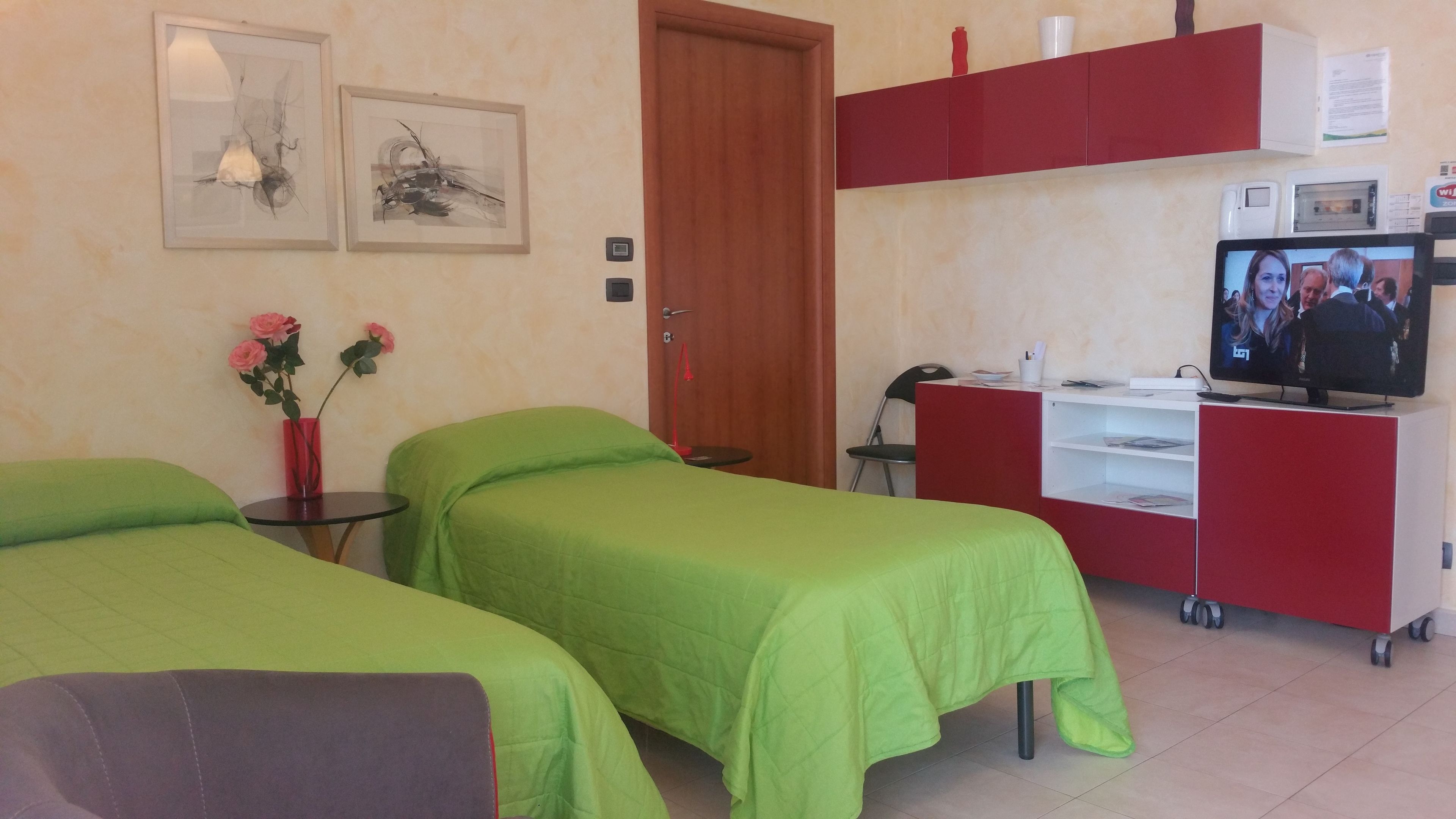 A hotel room with two single beds, a small table with a vase of flowers, a television on a stand, and a kitchenette area with a microwave and toaster.