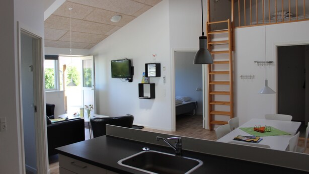 Una cocina moderna con una isla grande, un comedor con mesa y sillas, un dormitorio en lo alto con escalera, y un televisor montado en la pared.