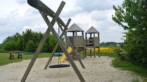 Un parque infantil con columpios y un tobogán, rodeado de arena y vegetación.