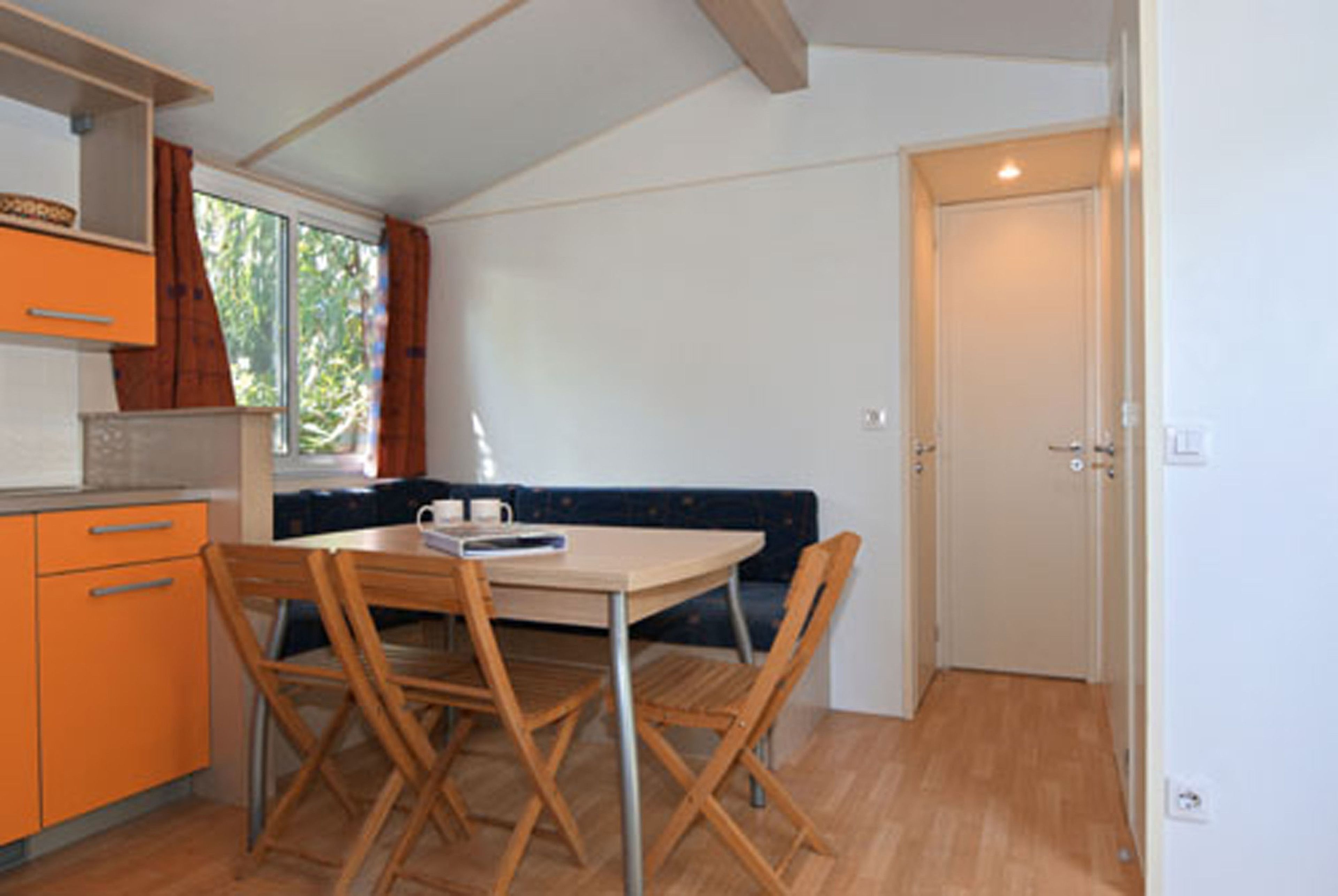 Een kleine keuken met een eettafel en stoelen, een raam met gordijnen, en een deur.