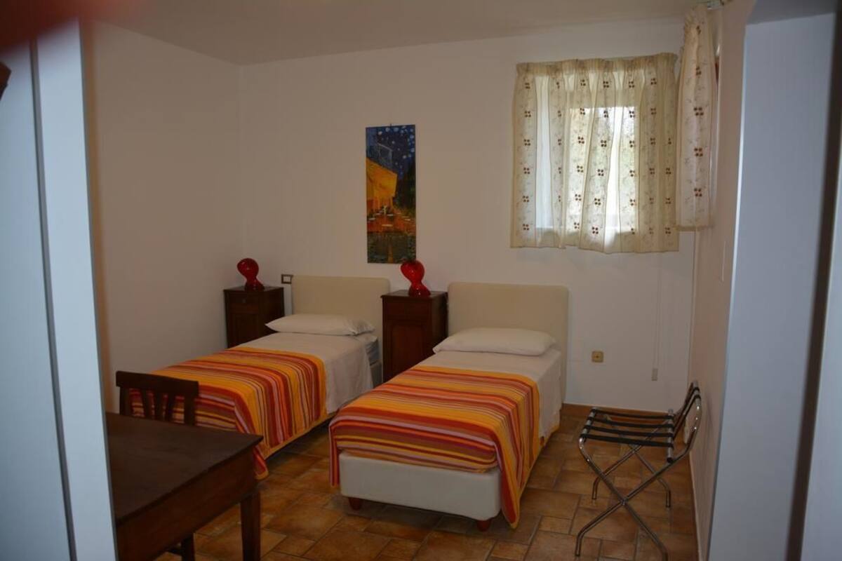 A small, clean room with two single beds, a small table, and a chair. There is a window with curtains and a painting on the wall.
