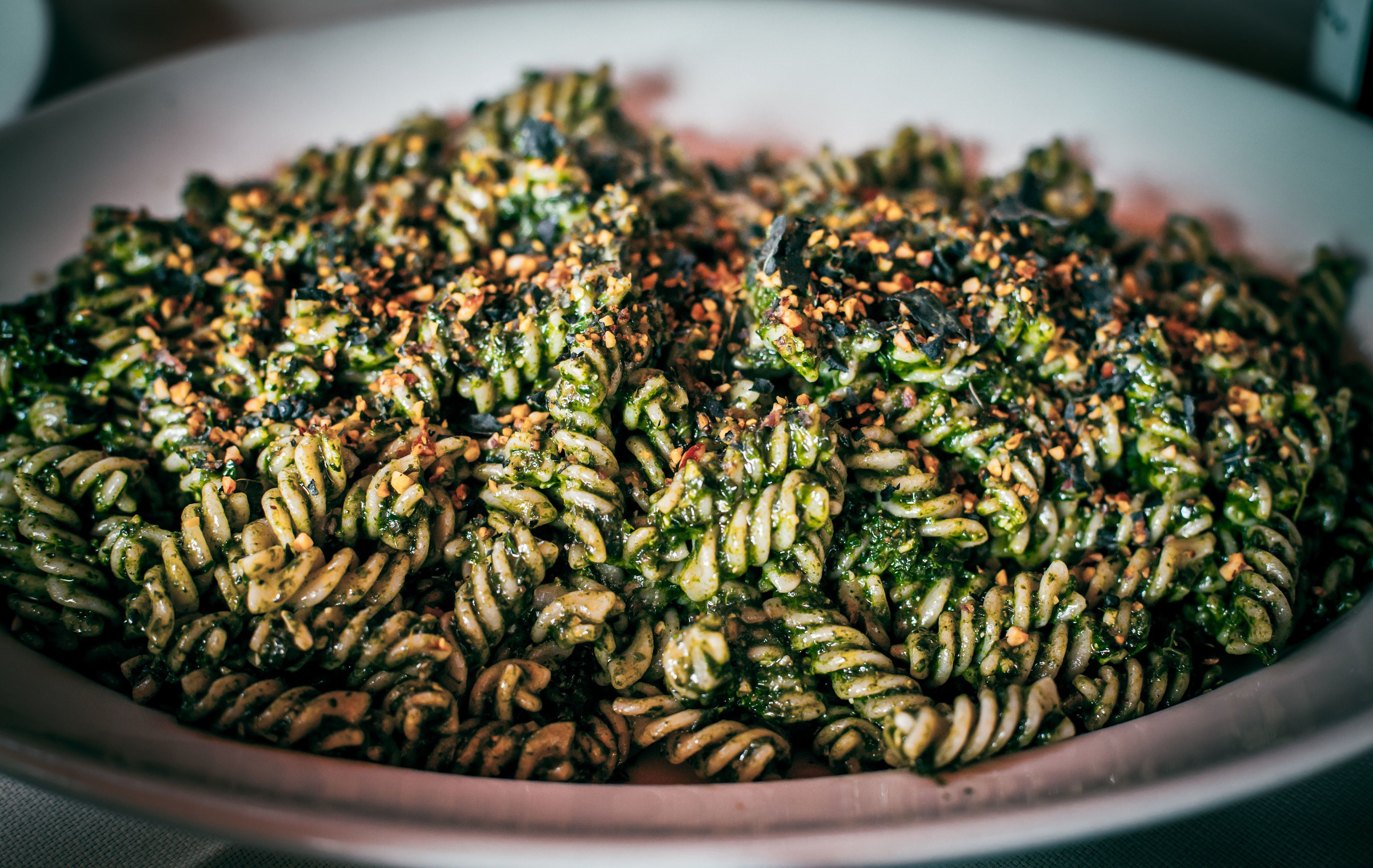 A large, shallow dish filled with a pasta dish, likely a pesto pasta, garnished with herbs and possibly nuts.