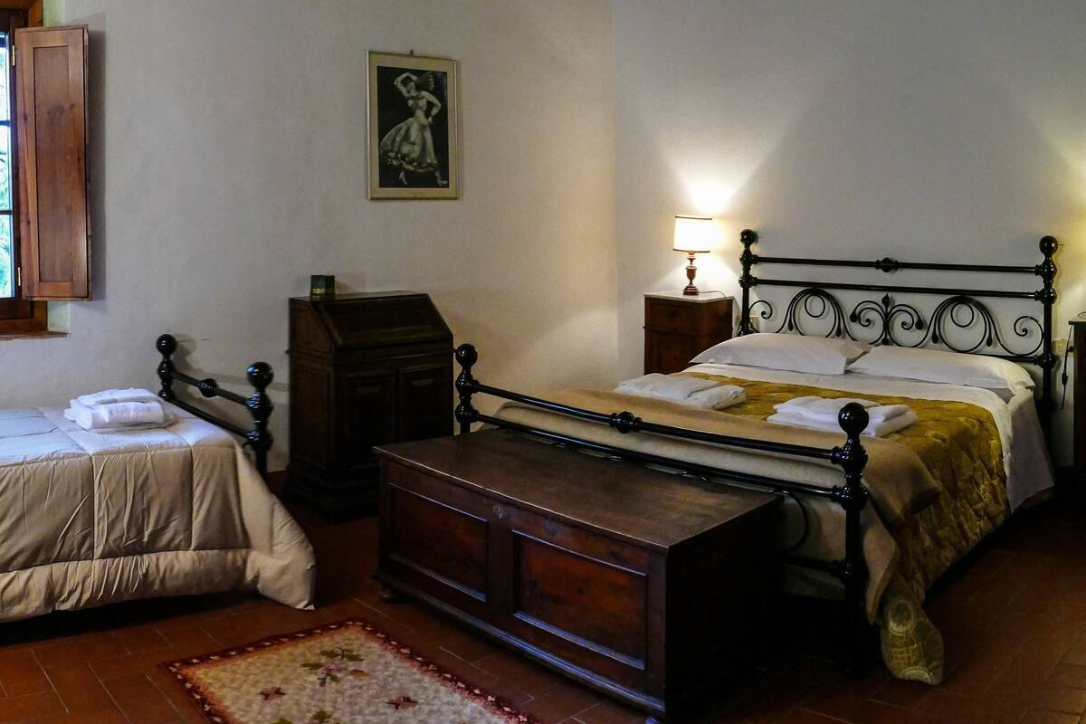 A traditional bedroom with two beds, a wooden dresser, a nightstand with a lamp, and a window with wooden shutters.