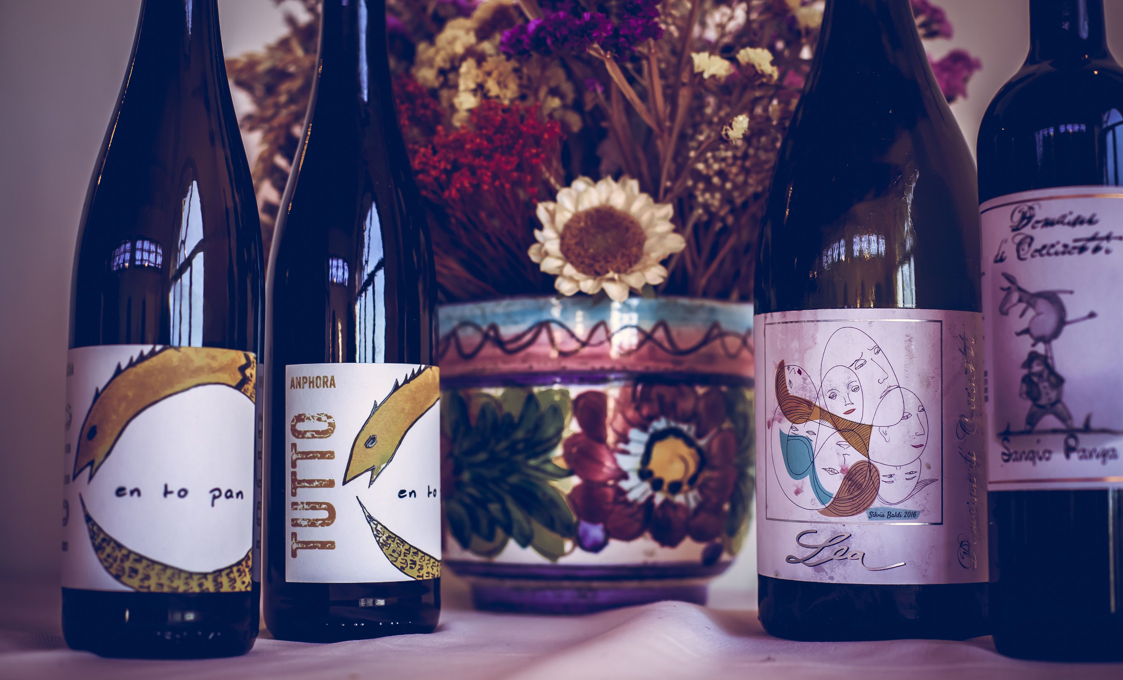 Cuatro botellas de vino con etiquetas distintivas, dispuestas sobre una mesa con un florero decorativo al fondo.