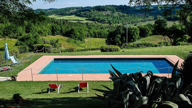 Une piscine rectangulaire entourée d’une clôture, deux chaises longues rouges et, en arrière-plan, une vue sur un paysage vallonné.