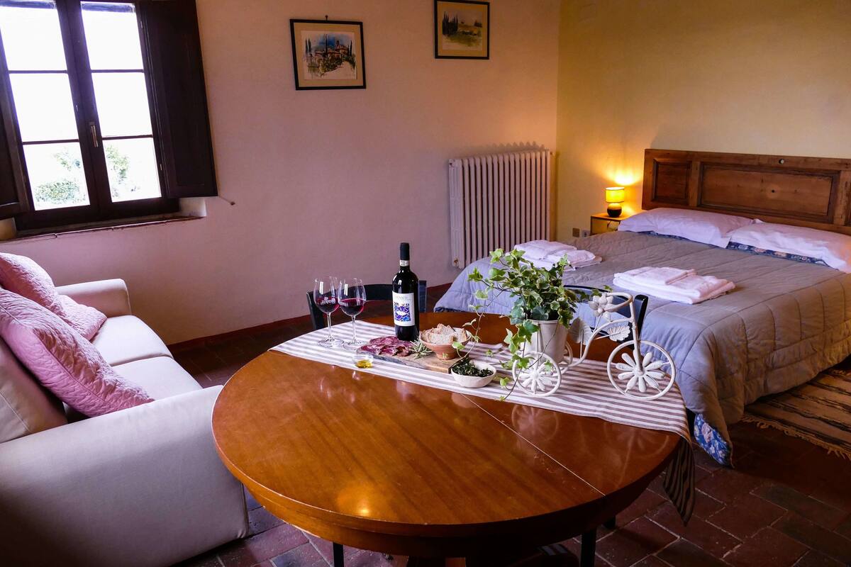 A hotel room with a bed, a dining table with a bottle of wine, glasses, and a plate of food.