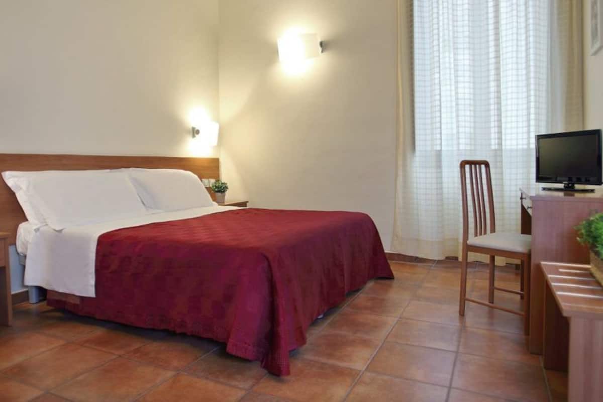 A hotel room with a bed, bedside tables, a desk with a chair, a television, and a window with curtains.