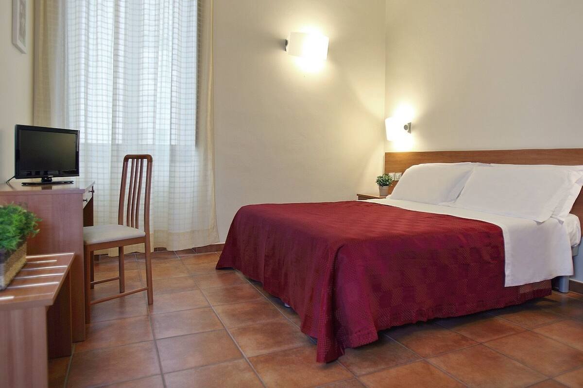A hotel room with a bed, a desk with a computer, a chair, and a small table with a plant.