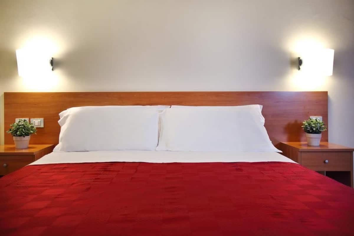 A neatly made bed with white linens and a red blanket, flanked by two bedside tables with potted plants.