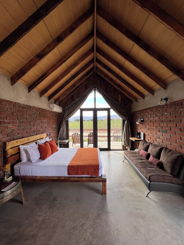 A bedroom with a wooden ceiling, brick walls, a bed with pillows, a sofa, and a view through large windows.