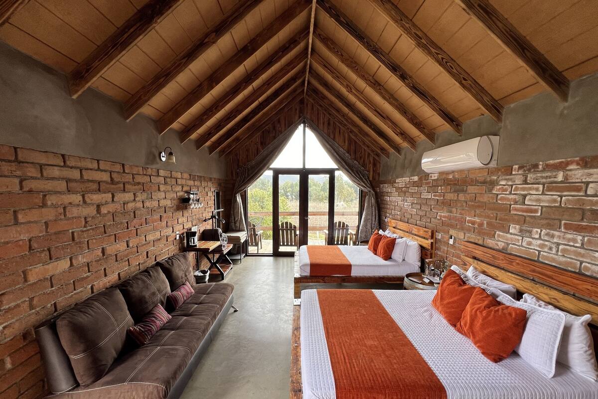 A rustic room with a high ceiling, brick walls, a wooden bench, a sofa, and two beds with orange pillows.