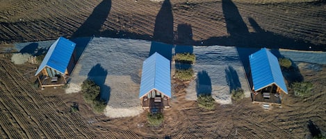 An aerial view of a property with three cabins, each with a blue roof, situated on a dirt road with a water body in the background.
