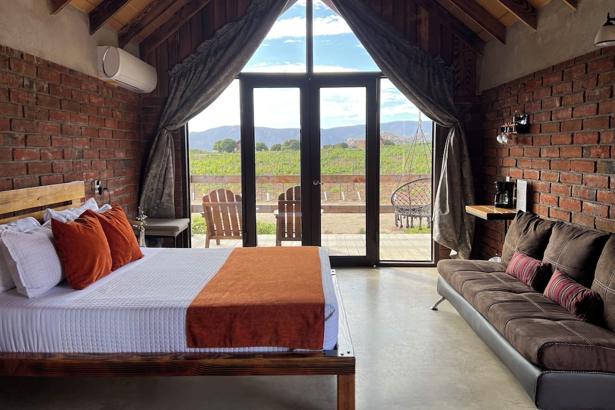 A bedroom with a bed, a wooden headboard, a brown sofa, and a view of a vineyard through large windows.