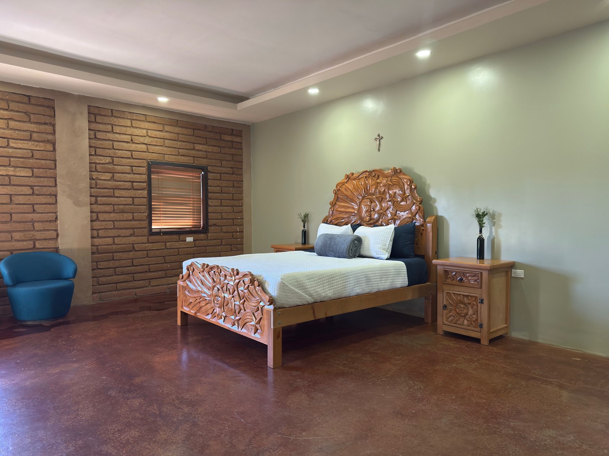 Quarto com cama de madeira, uma cadeira azul, parede de tijolos e uma pequena mesa de cabeceira de madeira.