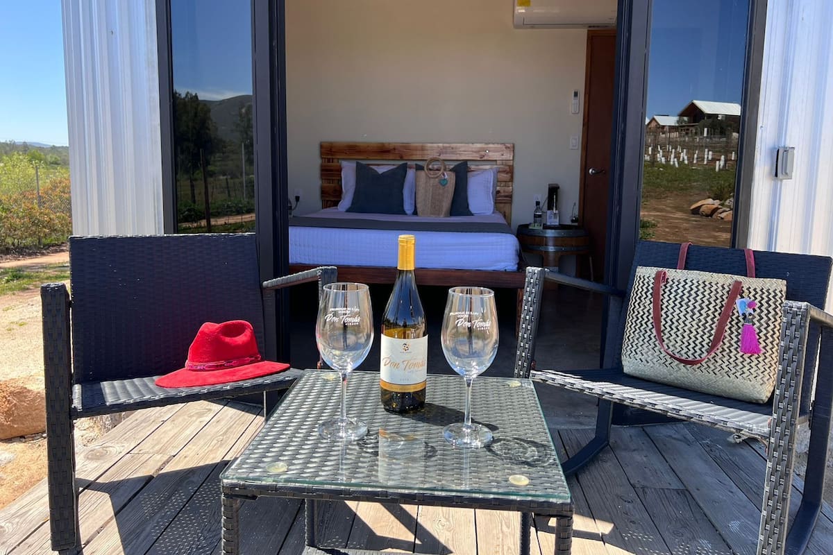 An open-air bedroom with a bed, a table with two wine glasses and a bottle, and a red hat on a chair.
