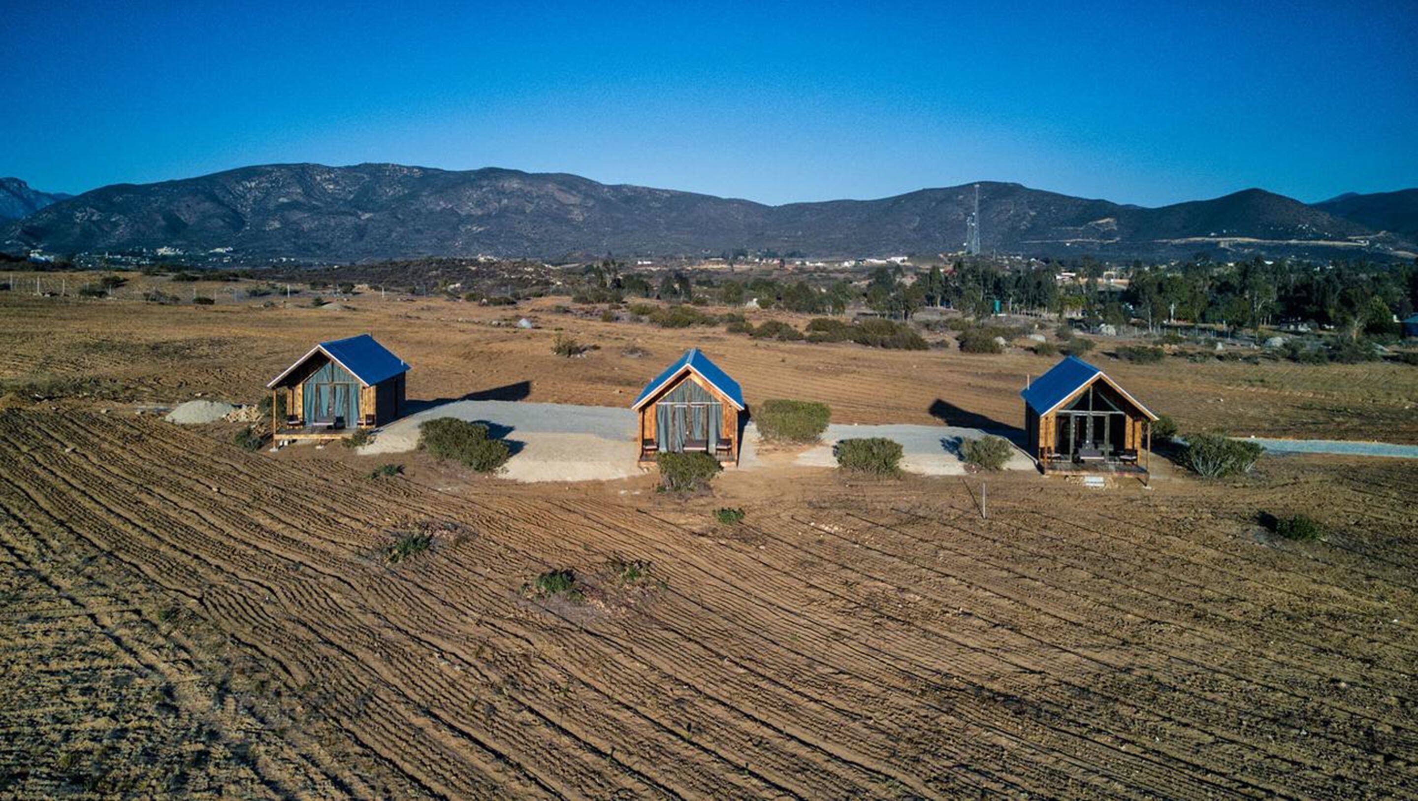 Três cabanas em um campo aberto e seco, com montanhas ao fundo.