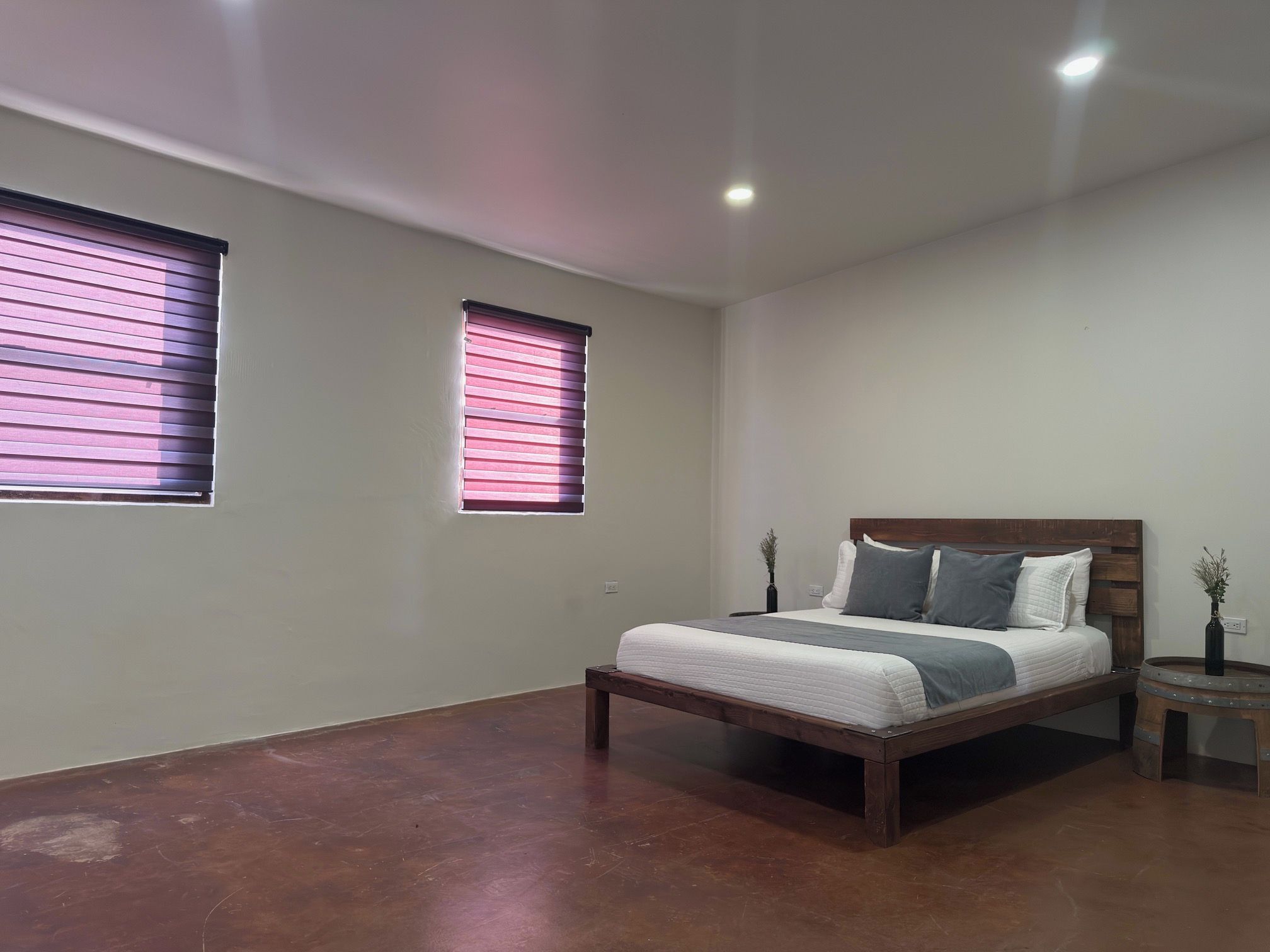 A bedroom with a bed, two windows with blinds, a bedside table, and a nightstand.