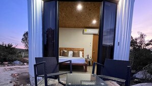 A circular building with a glass door leading to a bedroom with a bed, bedside table, and an air conditioning unit. There are two chairs and a small table on a wooden deck outside. - Don Tomas Viñedo Cabañas (Valle de Guadalupe)