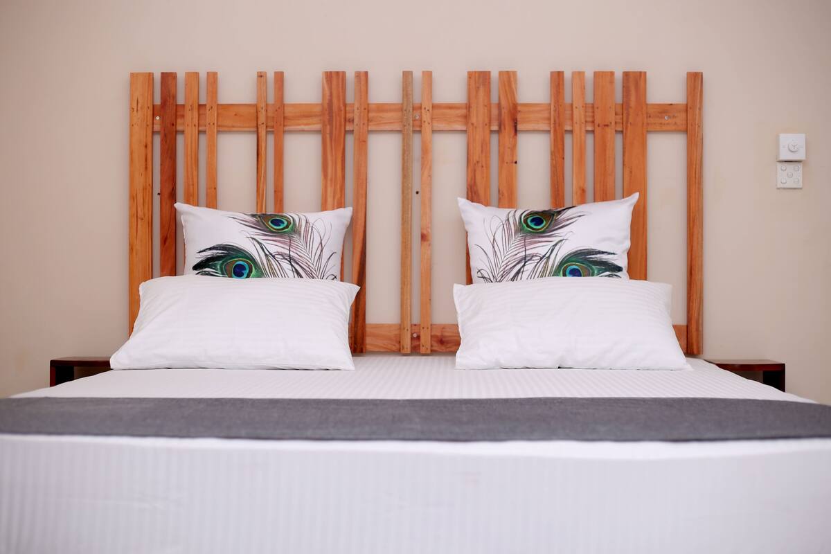 Un lit avec une tête de lit en bois, deux oreillers blancs ornés d’un motif de plumes de paon et un plaid gris.