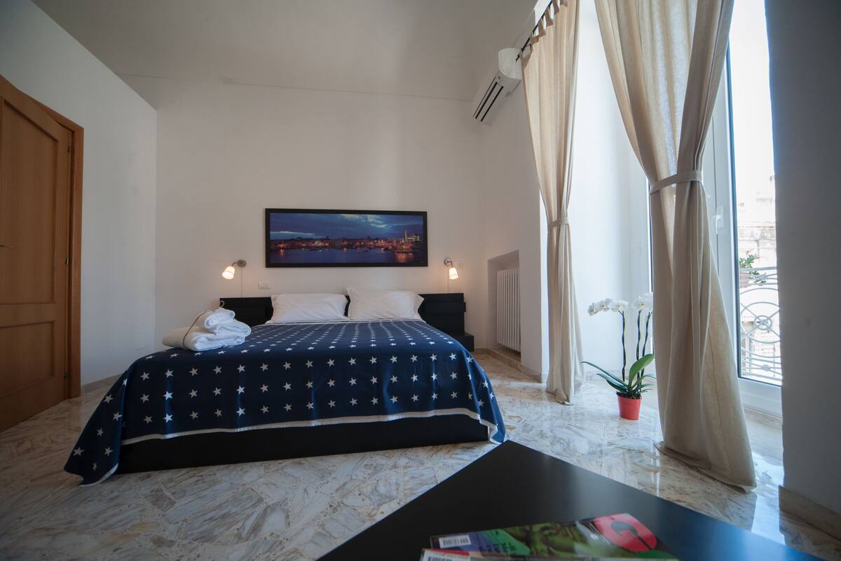 Un dormitorio con cama, mesita de noche, ventana con cortinas, planta en maceta y un cuadro en la pared.