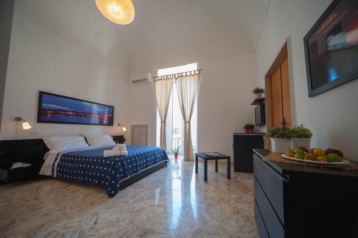 Un dormitorio con cama, mesita de noche, escritorio, cómoda, ventana con cortinas, televisor de pared y un cuadro enmarcado.