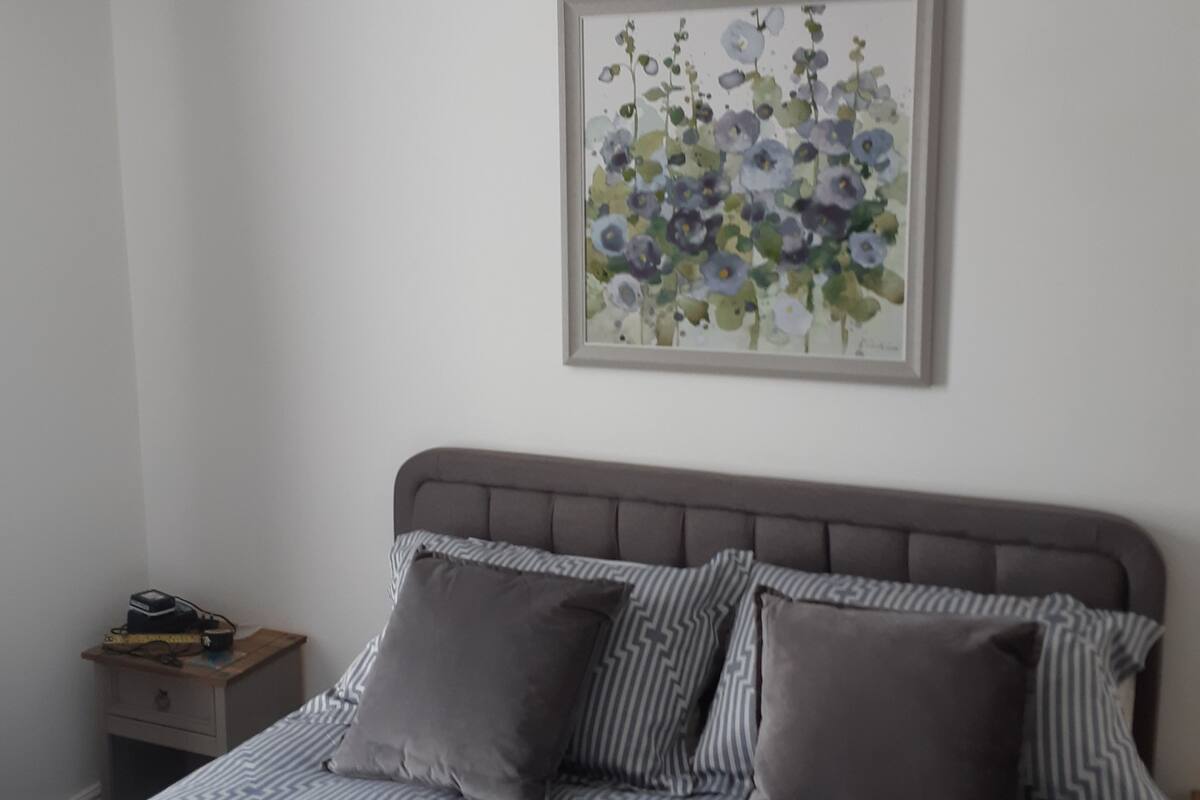 A bedroom with a bed, two bedside tables, a nightstand, a framed floral artwork, and two hanging lamps.