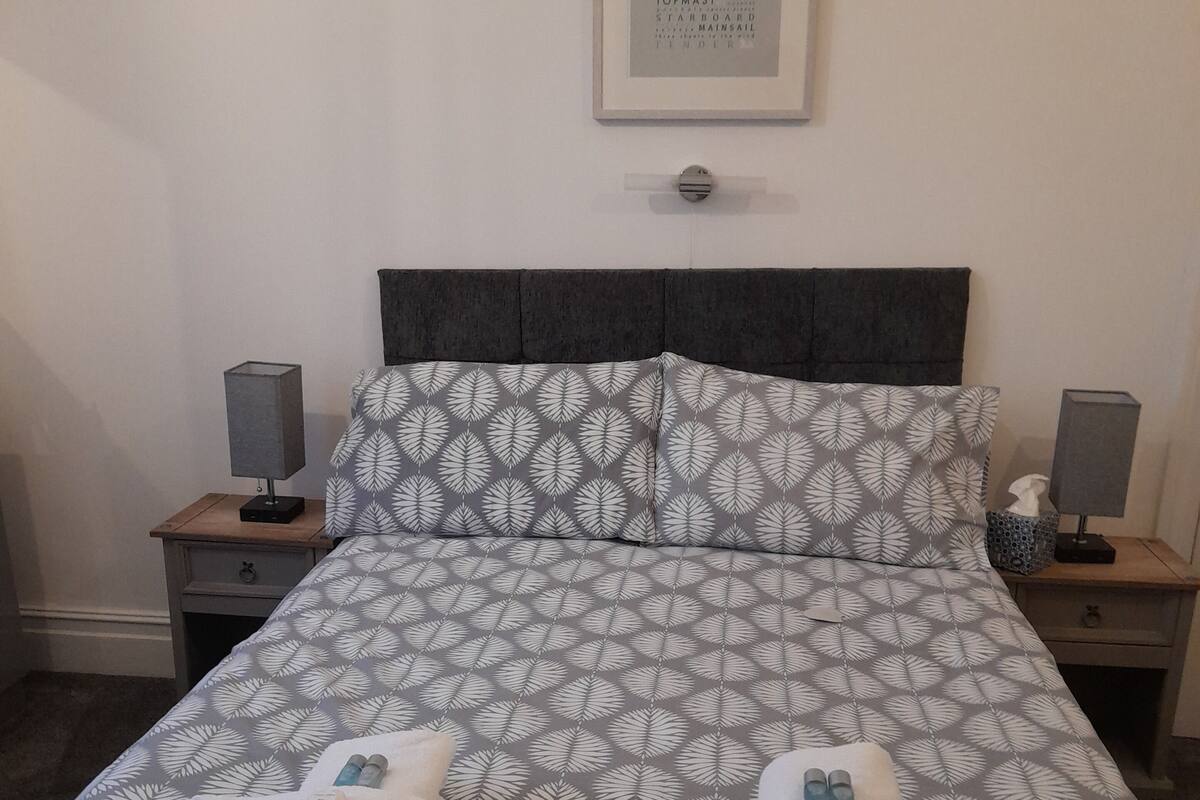 A neatly made bed with a leaf-patterned duvet, two towels, and a framed word puzzle on the wall.